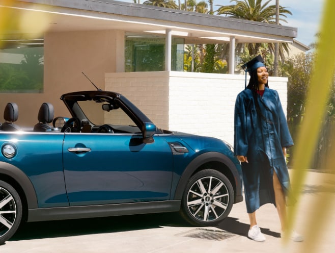 A girl in a graduation ensemble smiles by her MINI.